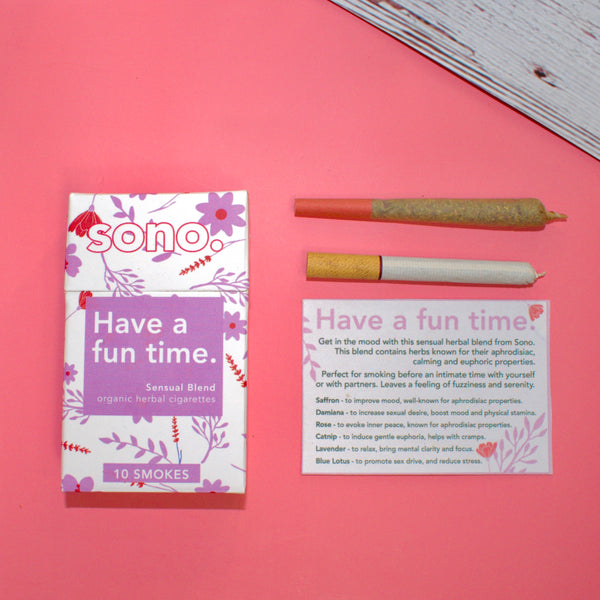 Image Description: A smoke pack lies next to its info card, 1 preroll & 1 cigarette, titled Have a Fun Time, an herbal aphrodisiac smoking blend. The smoke box is white & made with recycled paper and eco-friendly materials. There are pink designs on the white box, silhouettes of the herbs and flowers which are listed on the ingredient list: Damiana, Rose, Catnip, Lavender, Blue Lotus, and Saffron. Herbal smokes are also known as Herbal Spliffs, Herbal Cigarettes, Herbs & Flowers Prerolls, or Herb Tokes.