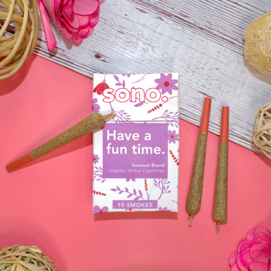 Image Description: A cardboard smoke pack titled Have a Fun Time, referring to the herbal aphrodisiac smoking blend that the 10 smokes inside are filled with. Beneath the title are the words Sensual Blends, & Organic Herbal Smokes. The box is decorated with pink floral shapes designed to look like the ingredients inside: Damiana, Rose, Catnip, Lavender, Blue Lotus, and Saffron. With the pack are 3 herbal prerolls, also known as Herbal Spliffs, Herbal Cigarettes, Herbs & Flowers Smokes, or Herb Tokes.